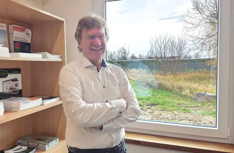 Ein lächelnder Mann im weißen Hemd steht mit verschränkten Armen in einem Büro neben einem großen Fenster, im Hintergrund sind ein Holzregal mit Informationsbroschüren und ein Blick ins Grüne zu sehen.