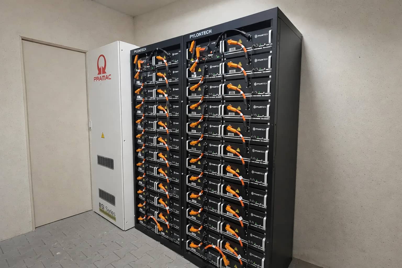 Gewerblicher Energiespeicher in einem Technikraum: Ein großes, schwarzes Pylontech Batteriesystem-Doppelrack mit zahlreichen Akkumodulen und orangefarbenen Kabeln steht neben einem weißen Pramac Steuerungsschrank der BSI Series.