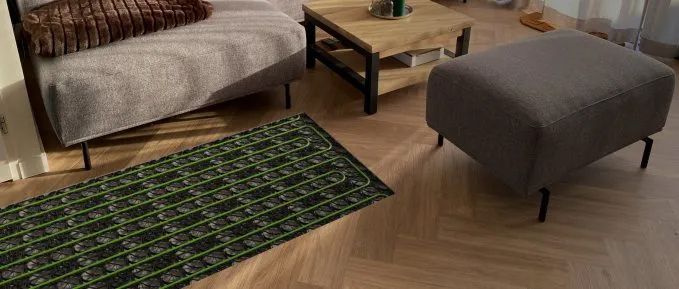 Wohnzimmer mit Fischgrätparkett, Couch, Couchtisch und einem Hocker, bei dem exemplarisch ein Bereich des Bodens freigelegt ist, um die darunter verlegten grünen Rohre einer Fußbodenheizung zu zeigen.